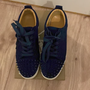 Mörkblå mocka sneakers med nitar - Säljer ett par mörkblå sneakers från Christian Louboutin i mocka med coola nitar på tån. Skorna har snörning, vit platt sula och den klassiska röda undersidan. Insidan är beige och de har en rund tå. Perfekt för dig som gillar edgy detaljer och lyxig stil.