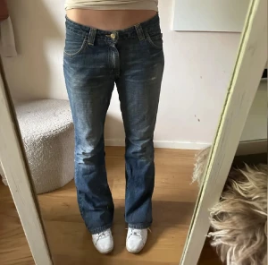 Blå bootcut jeans från Lee - vintage, lite slitna i grenen. Snygga blå jeans från Lee med klassisk bootcut passform och låg midja. Jeansen har fem fickor, slitna detaljer och Lee-logga på bakfickan. Tillverkade i robust denim med dragkedja och knapp framtill. Perfekta för en avslappnad och cool stil.