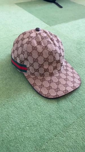 Gucci keps - Snygg beige keps från Gucci med klassiskt GG-mönster över hela kepsen. På sidan finns en ikonisk röd och grön rand. Justerbar rem baktill i mörkbrunt. Perfekt för dig som vill ha en lyxig streetstyle-accessoar. Finns i Stockholm kan även fraktas 