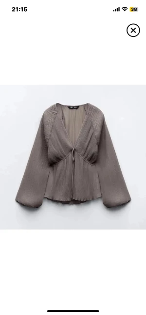 Grå omlottblus med knyt från ZARA - Säljer en snygg grå omlottblus från ZARA med plisserat tyg och v-ringning. Blusen har långa ärmar, knyt framtill och ett flowigt fall som ger en chill vibe. Perfekt att styla med jeans eller kjol för en cool look.
