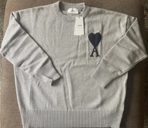 Grå stickad tröja från Ami Paris - Snygg grå stickad tröja från Ami Paris med svart hjärta och A-broderi på bröstet. Tröjan har rund hals, ribbade muddar och nederkant samt långärmad passform. Perfekt för dig som gillar stilrena detaljer och fransk design.