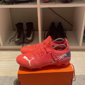 Röda Puma Ultra fotbollsskor - Säljer ett par snygga Puma Ultra fotbollsskor i röd färg med vita detaljer och logga. Skorna har snörning och en låg profil, tillverkade i syntetmaterial för lätt vikt och bra grepp på planen. Perfekt för dig som vill sticka ut med stil på fotbollen.
