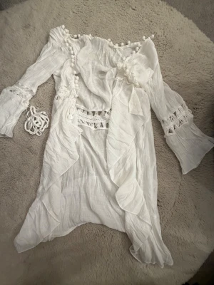 Vit cover - up  med volanger - Superfin vit omlottklänning med volangdetaljer och virkade partier i midjan och på ärmarna. Klänningen har långa ärmar med utsvängda avslut, dekorativa bollar längs halsringningen och medföljande knytband. Perfekt för sommarens alla tillfällen!