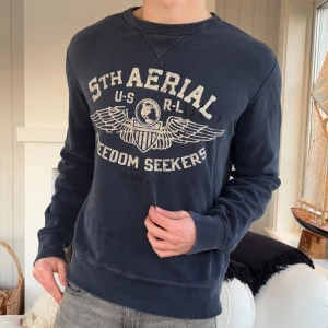 Mörkblå sweatshirt från Polo Ralph Lauren - Snygg mörkblå sweatshirt från Polo Ralph Lauren med tryck i vitt på bröstet och coolt vinge-motiv. Tröjan har rund hals, långa ärmar och ribbade muddar vid ärmslut och nederkant. Perfekt för dig som gillar streetwear och klassisk college-stil.