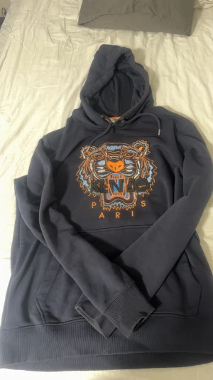 Mörkblå Kenzo hoodie med tigertryck - Säljer en mörkblå hoodie från Kenzo med ikoniskt broderat tigerhuvud i orange, blått och svart på bröstet. Tröjan har huva med snörning, ribbade muddar och magficka. Perfekt för dig som vill sticka ut med ett coolt streetwear-plagg.