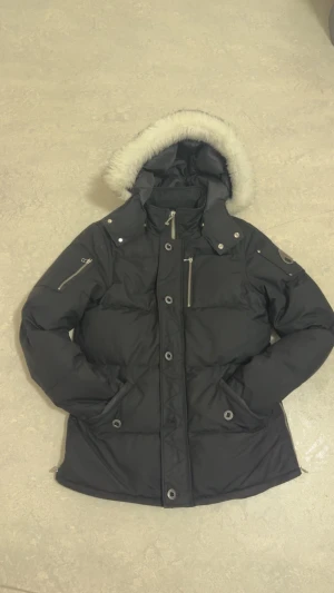 Svart dunjacka med päls på huvan - Svart dunjacka från Moose Knuckles med fluffig vit päls runt huvan. Jackan har flera dragkedjor, knappar och praktiska fickor framtill. Den är quiltad och har långärmade ärmar med dragkedjor på sidan. Perfekt för kalla vinterdagar.