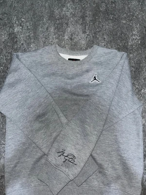 Grå Jordan sweatshirt med logga - Snygg grå sweatshirt från Jordan med klassisk Jumpman-logga broderad på bröstet och Michael Jordans signatur på ärmen. Tröjan har rund hals och långa ärmar, tillverkad i mjuk bomullsmix som är perfekt för chill eller sport. Enkelt stilren och riktigt skön.