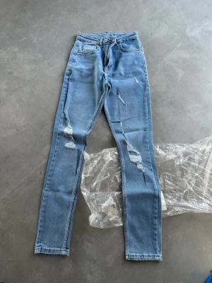 Nya Ljusa skinny jeans med slitningar XXS - Säljer ett par ljusblå skinny jeans i storlek XXS med coola slitningar på benen. Jeansen har klassisk femficksdesign, dragkedja och knapp framtill. Materialet är 98% bomull och 2% elastan för en skön stretchig passform.