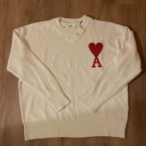 Vit stickad tröja från Ami - Vit stickad tröja från Ami Alexandre Mattiussi med röd hjärtlogga och bokstaven A på bröstet. Tröjan har rund hals, ribbade muddar och nederkant samt långärmad passform. Perfekt för dig som gillar clean design med en twist.