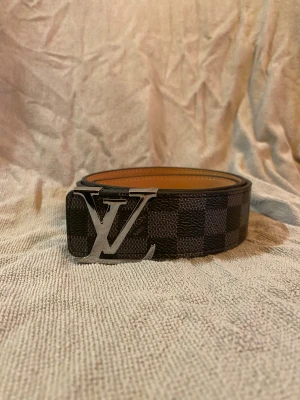 Louis Vuitton rutig svart skinnbälte - Säljer ett Louis Vuitton bälte i svart och grått med klassiskt rutigt mönster och stor LV-logga i silver på spännet. Bältet är gjort i skinn med beige insida och har en lyxig vibe som verkligen sticker ut. Perfekt accessoar för dig som gillar designer och vill ha något extra.