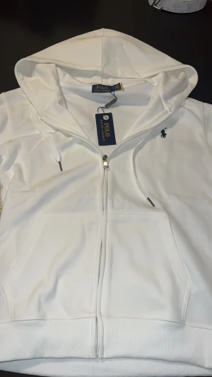 Vit hoodie från Polo Ralph Lauren - Vit hoodie från Polo Ralph Lauren med dragkedja och huva. Klassisk design med logobroderi på bröstet, två fickor fram och snörning i huvan. Tillverkad i mjuk bomullsmix som känns skön mot huden. Perfekt för chill dagar och streetwear.