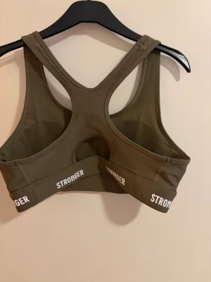 Olivgrön sport-bh från Stronger XL - Olivgrön sport-bh från Stronger i storlek XL med vita logotryck runt resåren och vita prickdetaljer på axlarna. Tillverkad av 75% återvunnen polyester och 25% elastan, har brottarrygg och bred resår för extra stöd. Perfekt för träning och aktiva dagar.