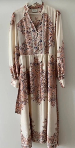  Långklänning från Neo Noir - Snygg långklänning från Neo Noir i ljusbeige med blommigt paisleymönster i rosa, blått och brunt. Klänningen har långärmade puffärmar, knappar framtill, markerad midja med bälte och lätt rynkad krage. Underbart satin-tyg!
