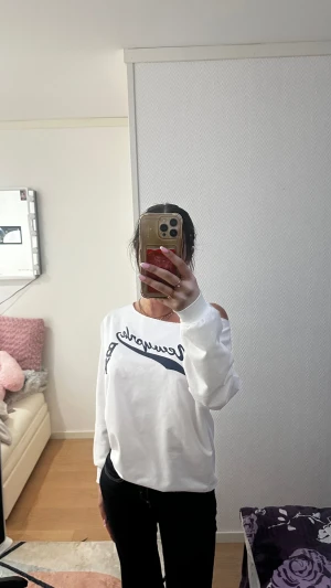 Vit offshoulder tröja  - Säljer en vit offshoulder tröja från Newyorkla med blått tryck framtill. Tröjan har lång ärm och en loose passform som ger en chill vibe. Materialet känns som mjuk bomull och den har en snygg öppen axel-design som gör den unik.