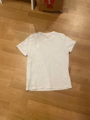 Vit basic t-shirt i bomull - En enkel vit t-shirt med korta ärmar och rund hals. Modellen är klassisk och passar perfekt till jeans eller shorts. Materialet är mjukt och skönt bomull, vilket gör den bekväm att bära hela dagen. Perfekt för dig som gillar en clean och stilren look.