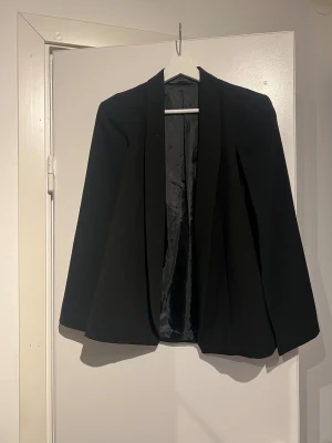 Svart kavaj med cape-ärmar från Dilvin - Svart kavaj från Dilvin Woman med cape-ärmar som ger en snygg och unik siluett. Jackan har en klassisk krage och är helfodrad med slät insida. Perfekt för dig som vill sticka ut med en stilren look. 