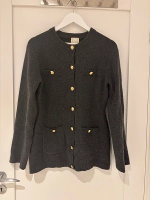 Mörkgrå kofta med guldknappar H&M - Snygg mörkgrå kofta från H&M med guldfärgade knappar framtill och på fickorna. Koftan har rund hals, två bröstfickor och två fickor nedtill. Stickad i mjukt material och har långa ärmar. Perfekt för dig som gillar klassiska detaljer.