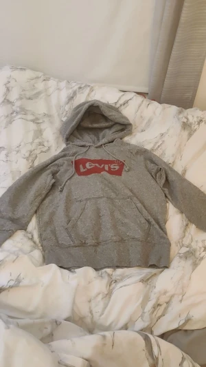 Jätte snygg Levis hoodie  - Säljer en grå hoodie från Levi's i storlek XS. Tröjan har en stor röd Levi's-logga på bröstet, huva med snörning och en klassisk magficka. Perfekt för chill dagar och enkel att matcha med jeans eller joggers.