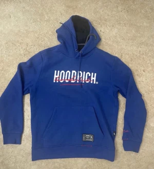 Blå hoodie från Hoodrich med tryck - Snygg blå hoodie från Hoodrich med vit och röd logga på bröstet och ryggen. Tröjan har huva med snörning, känguruficka och ribbade muddar. Detaljer med rött tryck på ärmen och nedtill. Perfekt för dig som gillar streetwear och vill sticka ut.