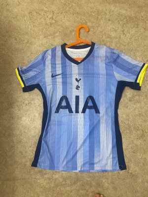 Tottenham Hotspur blå randig fotbollströja - Snygg Tottenham Hotspur fotbollströja från Nike i olika blå nyanser med vertikala ränder. Kortärmad modell med mörkblå V-ringad krage och gula detaljer vid ärmslut. Materialet är lätt och andas, perfekt för träning eller match. Stor AIA-logga på bröstet.