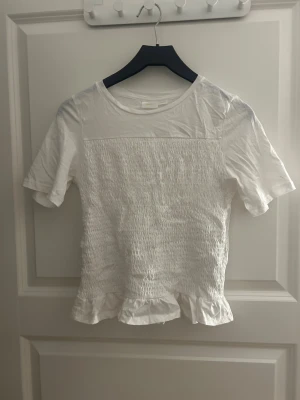 Vit blus med volangkant - Säljer en söt vit blus med korta ärmar och rund hals. Toppen har smockad framsida som ger en cool textur och avslutas med en volangkant nedtill. 