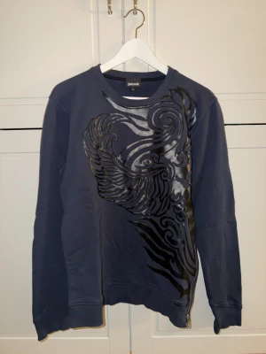 Blå sweatshirt från Just Cavalli - Snygg mörkblå sweatshirt från Just Cavalli med ett stort, glansigt svart mönster framtill. Tröjan har rund hals, långa ärmar och en cool dragkedja vid nederkanten. Perfekt för dig som gillar statement-plagg och vill sticka ut.