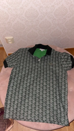 Mönstrad piké från Lacoste - Snygg pikétröja från Lacoste med geometriskt mönster i svart och grått. Klassisk krage i svart, korta ärmar och den ikoniska krokodilloggan på bröstet. Insida i grön färg ger en cool kontrast. Perfekt för dig som gillar street och sportig stil. Använd när jag va utomlands i 2 veckor endast