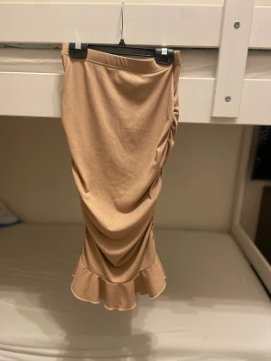 Beige långkjol med volangkant - Snygg beige långkjol i mjukt ribbat material med draperad effekt längs sidan och volangkant längst ner. Kjolen har en slim passform som framhäver siluetten och ger en cool vibe. Perfekt för dig som gillar stilrena och trendiga plagg med extra detaljer.