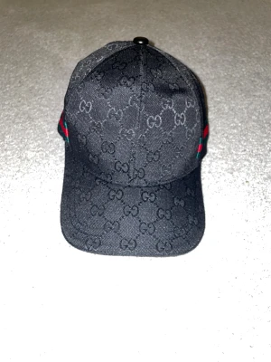 Svart keps med GG-mönster - Snygg svart keps från med GG-monogram över hela ytan. Kepsen har en böjd skärm, justerbar rem i skinn baktill och ikoniska gröna och röda ränder på sidan. Materialet är bomull och polyester, med exklusiva detaljer och lyxig känsla.