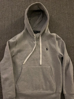 Grå hoodie från Polo Ralph Lauren - Snygg grå hoodie från Polo Ralph Lauren i custom fit. Tröjan har vit snörning vid huvan, klassisk känguruficka och det ikoniska Polo-logot broderat på bröstet. Perfekt för chill dagar och enkel att matcha med jeans eller joggers.
