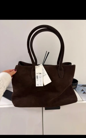 Mörkbrun mocka handväska från ARKET - Snygg och rymlig mörkbrun handväska från ARKET i mjuk mocka. Väskan har två rejäla handtag, magnetknäppning och en innerficka med dragkedja. Perfekt storlek för att få plats med allt du behöver och stilren design som passar till många outfits.