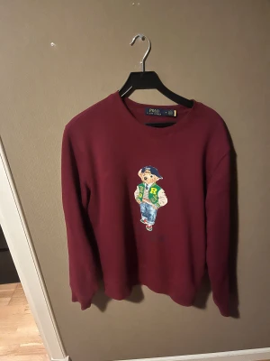 Vinröd sweatshirt Polo Ralph Lauren - Vinröd sweatshirt från Polo Ralph Lauren med klassisk björn-tryck på bröstet. Tröjan har rund halsringning och långa ärmar. Skön och stilren modell i mjuk bomull, perfekt för dig som gillar streetwear med en preppy twist.