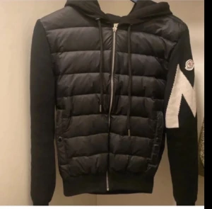 Svart Moncler pufferjacka med huva - Svart pufferjacka från Moncler med huva och dragkedja framtill. Jackan har quiltad framsida, stickade ärmar med vit zigzag-detalj och Moncler-logga på vänster ärm. Den är 100% äkta