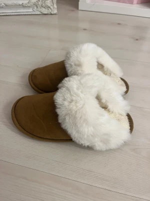 Bruna fluffiga tofflor med vit päls - Supermysiga bruna tofflor med vit fluffig päls runt kanten. Utsidan är i mocka-look och insidan är fodrad med mjuk päls för extra komfort. Perfekta att glida runt i hemma när det är kallt ute. Rund tå och platt sula för skön känsla.