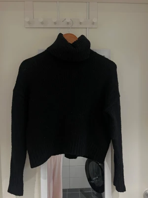 Svart stickad polotröja - Mysig svart stickad polotröja med ribbad krage och muddar. Tröjan har lång ärm och en loose passform som ger en chill vibe. Perfekt för kalla dagar när du vill vara både bekväm och snygg. Materialet är grovstickat och känns mjukt mot huden.