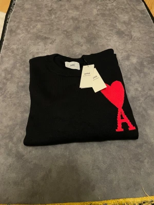 Svart stickad tröja från Ami Paris - Svart stickad tröja från Ami Paris med röd hjärtformad A-logga på bröstet. Tröjan har rund hals, ribbade muddar och är långärmad. Materialet är mjuk bomull och passformen är normal. Perfekt för dig som gillar stilrena plagg med en unik detalj.