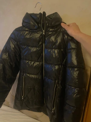 Svart pufferjacka från Everest - Säljer en svart pufferjacka från Everest med glansig finish och huva. Jackan har två stora fickor med dragkedja och tydliga quiltade sektioner. Perfekt för kalla dagar och har en snygg, sportig look. Everest-logga på ärmen och insidan.