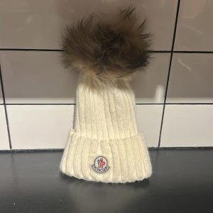 Vit Moncler mössa med pälsboll - Ribbstickad vit mössa från Moncler med stor fluffig pälsboll på toppen. Märkets klassiska logga framtill på uppvikt kant. Mössan är gjord i mjuk ull och har en lyxig vibe som sticker ut i vinterkylan.