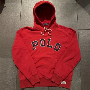 Röd hoodie från Polo Ralph Lauren - Säljer en röd hoodie från Polo Ralph Lauren med stor broderad POLO-text i blått och vitt på bröstet. Tröjan har huva med vit snörning, känguruficka och en patch med Polo RL-detaljer vid nederkanten. Perfekt för dig som gillar klassisk streetwear.