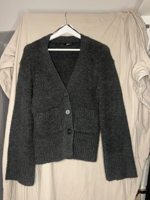 Mörkgrå stickad kofta från Gina Tricot - Mysig mörkgrå stickad kofta från Gina Tricot med v-ringad hals och tre knappar framtill. Koftan har två praktiska fickor och ribbade muddar vid ärmslut och nederkant. Perfekt för lager-på-lager och chill dagar.
