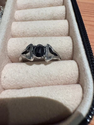 Silverring med svart sten - Snygg silverfärgad ring med en stor svart, fasettslipad sten i mitten och detaljerade mönster längs sidorna. Ringen har en unik design med öppna partier och små dekorationer som ger den en cool och mystisk vibe. Perfekt för dig som gillar statement-smycken.