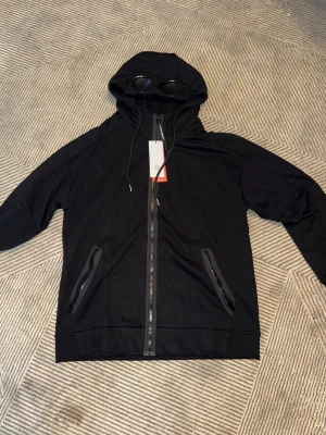 Svart vindjacka från C.P. Company - Svart vindjacka från C.P. Company med ikoniska glasögon i huvan. Jackan har hel dragkedja framtill, två fickor med dragkedja och justerbar huva med snörning. Perfekt för dig som gillar streetwear och vill sticka ut med detaljer.