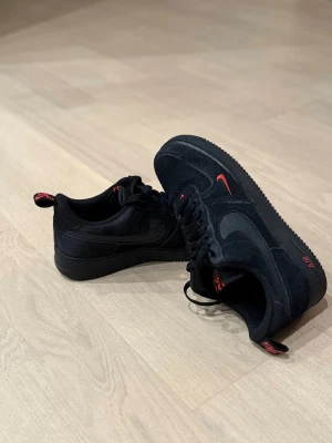 Nike Air Force 1 - Svarta Nike Air Force 1 sneakers med röda detaljer på logga, sula och snörning. Låg modell med klassisk rund tå och platt sula. Ovandel i mocka och syntet, snyggt broderad swoosh på sidan. Perfekt för dig som gillar stilrena och coola sneakers. Köpta från JD för ca ≈1500 och är fortfarande i ganska bra skick då de är använda ett fåtal ggr