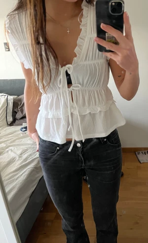 Vit blus med volanger och snörning - Supersöt vit blus med volanger och knyt framtill. Toppen har en v-ringad design. Materialet är lätt och luftigt, perfekt för varmare dagar. 