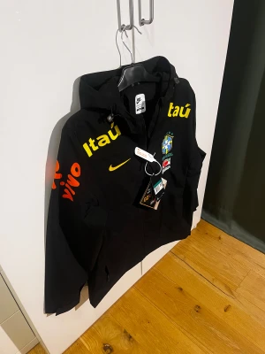 Svart Nike Brasil vindjacka XL/175~M - Nike Brasil vindjacka i svart med gula och orangea tryck, Brasil-logga på bröstet och huva. Jackan har dragkedja framtill och meshfoder på insidan. Snygga detaljer med sponsorer på ärmarna och bröstet. Perfekt för dig som gillar fotboll och streetwear. Står XL men är strl M(fabriksfel) men sitter som en M.