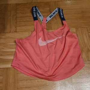 Nike Dri-Fit rosa linnetopp XS - Sportig rosa linnetopp från Nike med vita och marinblå axelband där det står 'JUST DO IT'. Toppen har ett vitt Nike-logga framtill och är tillverkad i Dri-Fit material som andas. Perfekt för träning och aktiva dagar.