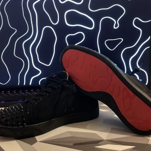 Christian Louboutin svarta sneakers med nitar - Säljer ett par svarta sneakers från Christian Louboutin med ikonisk röd sula och coola svarta nitar över tån. Skorna har snörning, rund tå och är tillverkade i skinn med textilfoder. Perfekt för dig som vill sticka ut med en edgy och lyxig stil.