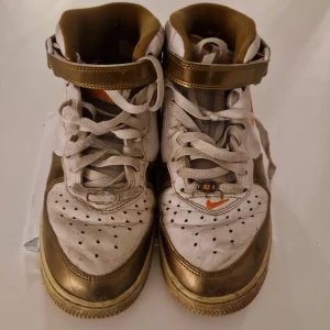 Nike Air Force 1 High guld/vit - Unika Nike Air Force 1 High sneakers i guld och vitt med orange swoosh och detaljer. Skorna har snörning, högt skaft med kardborreband och perforerad tå. Tillverkade i skinn med platt sula och rund tå. Perfekt för dig som vill sticka ut med en fet färgkombo.Den som köper betalar för frakt även om det ska till ett anat land 
