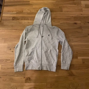 Grå hoodie från Polo Ralph Lauren - Snygg grå hoodie från Polo Ralph Lauren med dragkedja och huva. Tröjan har snörning vid huvan, två fickor framtill och den klassiska Polo-loggan broderad på bröstet. Perfekt för chill dagar och enkel att matcha med jeans