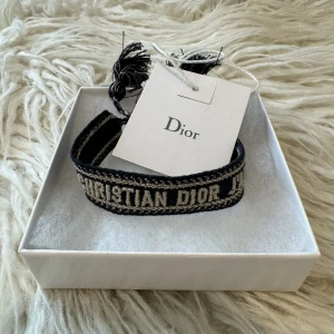 Christian Dior armband  -  Snyggt vävt armband från Dior i beige och marinblått med tofsdetaljer. Klassisk modell med texten “CHRISTIAN DIOR J’ADIOR”. Perfekt accessoar för både vardag och fest 